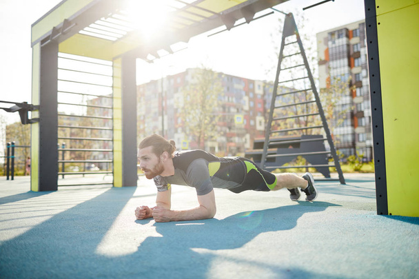 Serious young handsome man with red beard leaning on elbows while performing plank exercise on outdoor sports ground - Fotografie, Obrázek