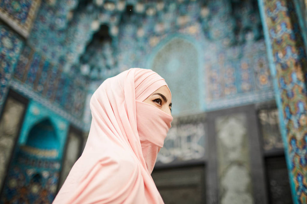 Senhora muçulmana enigmática com rosto coberto indo para a bela mesquita com design ornamental e olhando para longe
 - Foto, Imagem