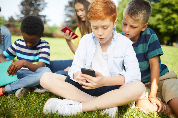 Dört sınıf arkadaşı, dışarıda çimenlerin üzerinde oturup telefonla oynayarak sınıflar arasında mola veriyor. - Fotoğraf, Görsel