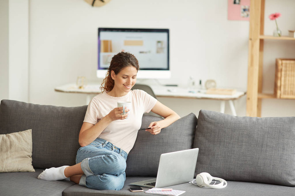 Positiivinen houkutteleva nuori nainen istuu mukavalla sohvalla ja juo kahvia tehdessään verkko-ostoksia
 - Valokuva, kuva