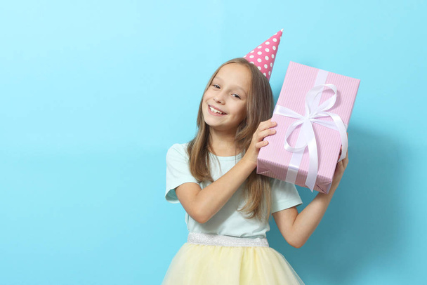 Portrait of a little girl in a festive cap and with a gift in her hands on a colored background. Birthday, presents, holiday - Фото, зображення