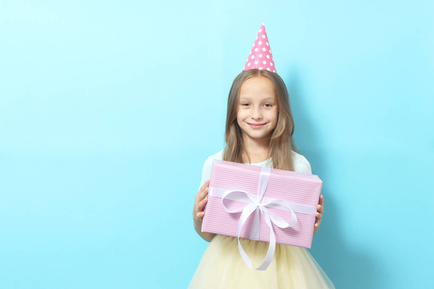 Portrait of a little girl in a festive cap and with a gift in her hands on a colored background. Birthday, presents, holiday - Zdjęcie, obraz