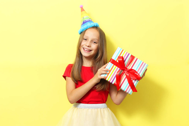 Porträt eines kleinen Mädchens mit festlicher Mütze und einem Geschenk in der Hand auf farbigem Hintergrund. Geburtstag, Geschenke, Urlaub - Foto, Bild