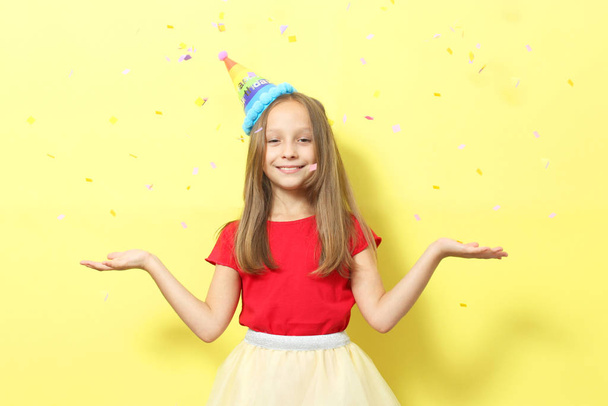 Portret van een klein meisje met een feestelijke pet en een cadeau in haar handen op een gekleurde achtergrond. Verjaardag, cadeautjes, vakantie - Foto, afbeelding