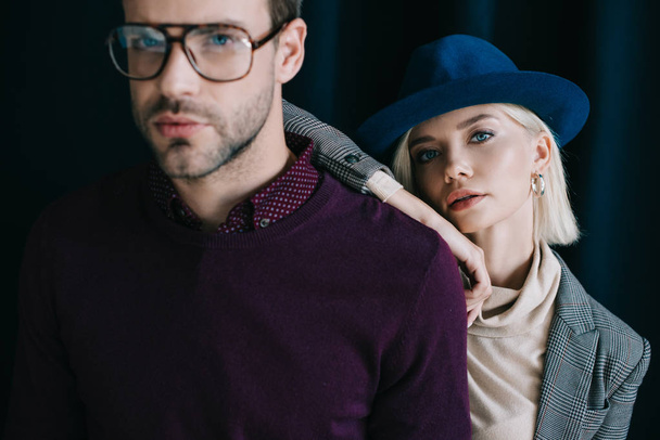 stijlvolle jonge man in glazen en blonde vrouw in hoed kijken naar camera in de buurt van gordijn - Foto, afbeelding