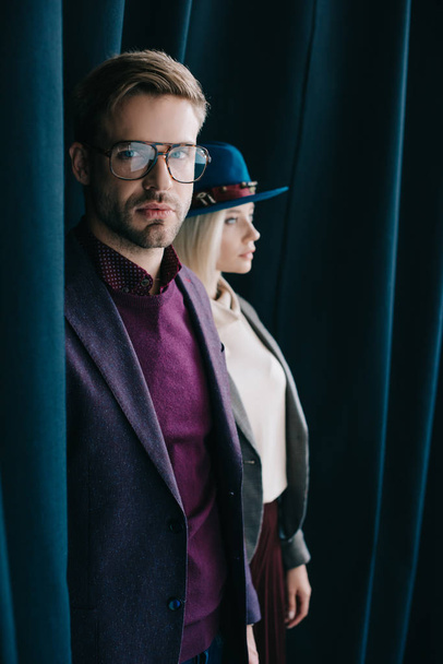stylish young man in glasses and blonde woman in hat near curtain - Fotó, kép