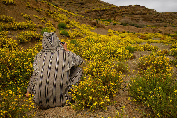 Widok z tyłu człowieka w stylu arabskim szaty siedząc i robienia zdjęć małe żółte kwiaty na wzgórzach gór Atlas, Maroko - Zdjęcie, obraz