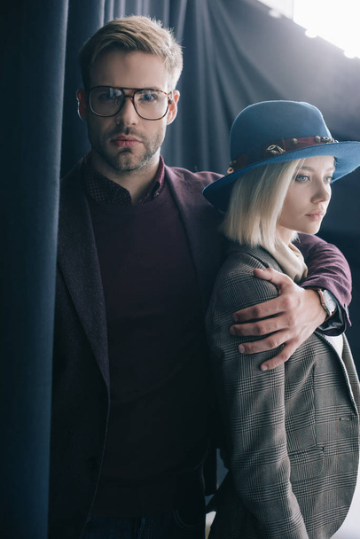 elegant young man in glasses embracing blonde girl in hat near curtain - Foto, afbeelding