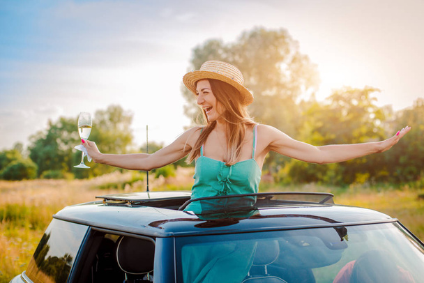 Radosna dziewczyna kobieta raduje się kieliszkiem wina wychylając się z włazu samochodu na imprezie w lecie w naturze. - Zdjęcie, obraz