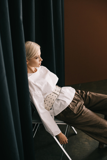 stylish blonde woman in white blouse sitting on chair near curtain on brown - 写真・画像
