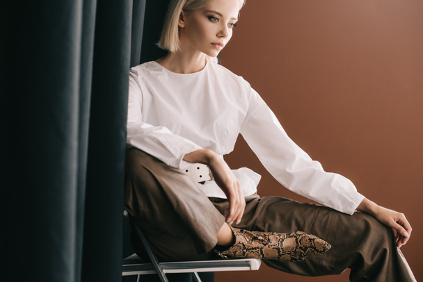 stylish blonde woman in white blouse sitting on chair near curtain on brown - Photo, Image