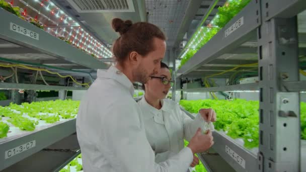 Δύο επιστήμονες, ένας άντρας και μια γυναίκα με άσπρα παλτά βάζουν δείγματα φυτών σε ένα δοκιμαστικό σωλήνα για να ανακαλύψουν τα αποτελέσματα των εξετάσεων. - Πλάνα, βίντεο