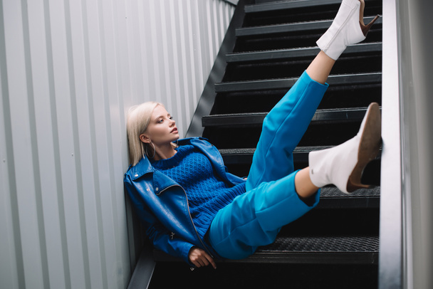 Blonde Frau in blauer Lederjacke und High Heels sitzt auf Treppe - Foto, Bild