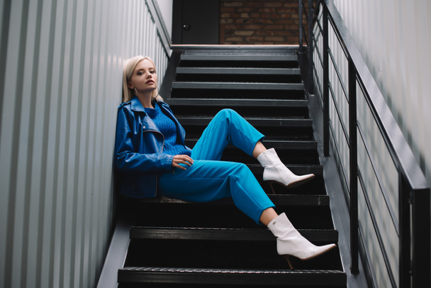 blonde woman wearing blue leather jacket and heels sitting on stairs - Foto, immagini
