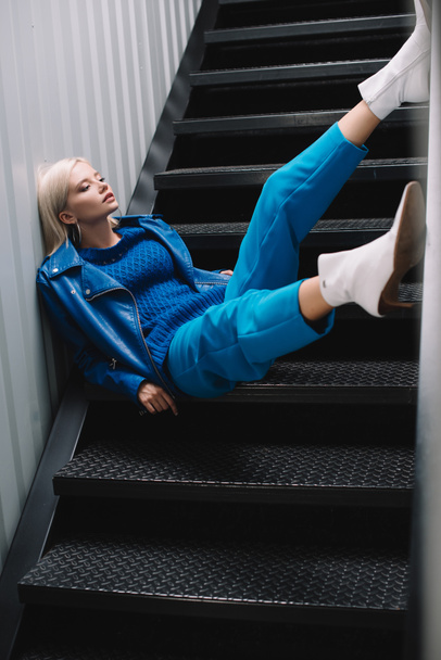 blonde woman wearing blue leather jacket and heels sitting on stairs - Foto, imagen