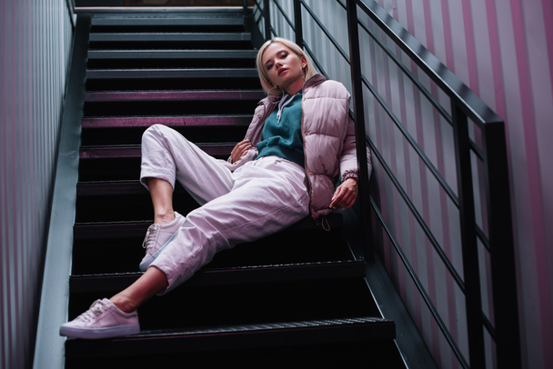 confident blonde girl in down jacket and sneakers sitting on stairs - Foto, Imagem