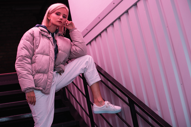 blonde young woman in down jacket looking at camera on stairs - Foto, Imagem