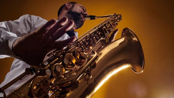 Portrait of professional musician saxophonist man in  white shirt plays jazz music on saxophone, yellow background in a photo studio, bottom view - Zdjęcie, obraz