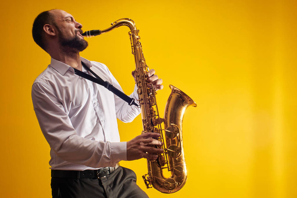 Portrait of professional musician saxophonist man in  white shirt plays jazz music on saxophone, yellow background in a photo studio, side view - Photo, Image