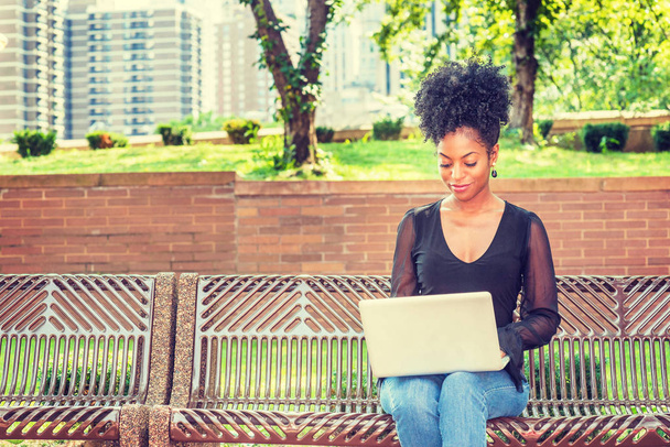 メッシュの薄い長袖シャツ、ジーンズを着たアフロヘアスタイルの若いアフリカ系アメリカ人女性、ニューヨークの公園のベンチに座って、下を見て、読書、ラップトップコンピュータで働く - 写真・画像