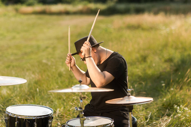 A drummer in glasses plays drums in the countryside. The man has a black hat, behind him an old castle - Photo, Image