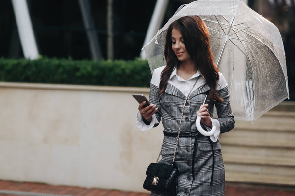 Retrato de una hermosa mujer en anticipación de conocer a su novio. Mujer con abrigo gris bajo el paraguas, sosteniendo el teléfono inteligente en la mano. Es lluvia primaveral
. - Foto, Imagen