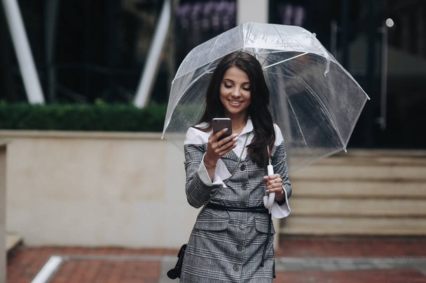 Horizontal portrait of beautiful smiling woman with smartphone in hand and reading sms. Woman wearing gray coat under umbrella - Фото, зображення