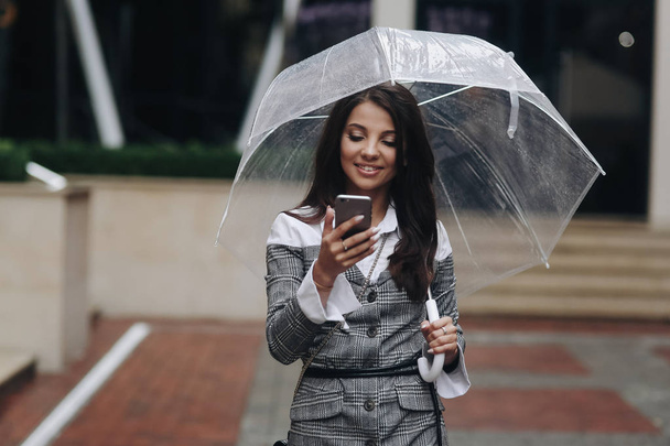 Portrait of beautiful smiling woman with smartphone in hand and typing sms. Woman wearing gray coat under umbrella received a funny SMS message - Foto, Imagen