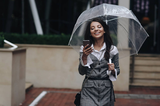 Portrait of beautiful smiling woman with smartphone in hand and typing sms standing near cafe. Woman wearing gray coat under umbrella - Фото, изображение