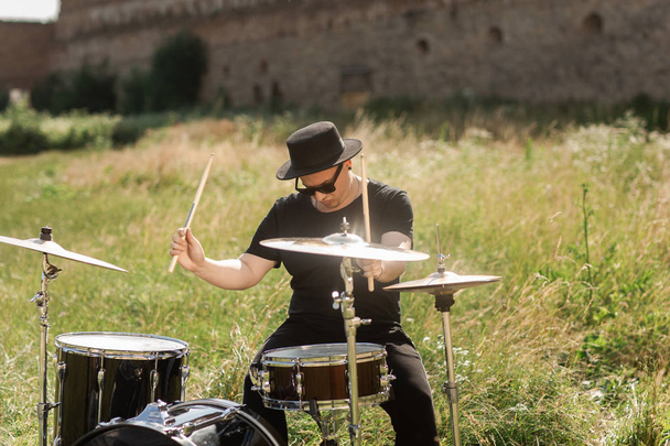 A drummer in glasses plays drums in the countryside. The man has a black hat, behind him an old castle - Photo, Image