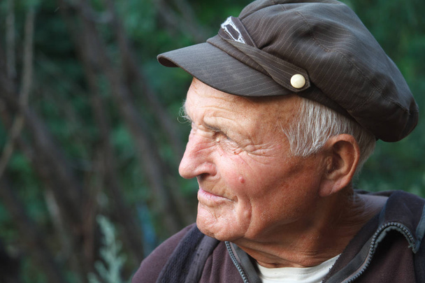 Close-up portrait of a very old man in a cap on a blurred background of green trees, selective focus - Photo, Image