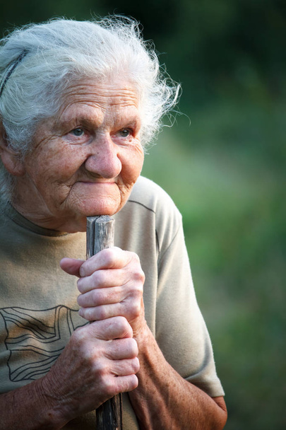 Un retrato de cerca de una anciana con el pelo gris sonriendo y mirando hacia arriba, apoyando su barbilla en un palo como si caminara con un bastón, cara en arrugas profundas, enfoque selectivo
 - Foto, Imagen