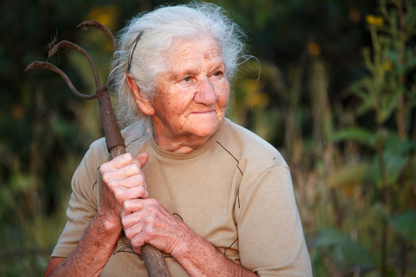 Κοντινό-up πορτρέτο μιας παλιάς γυναίκας με γκρίζα μαλλιά κρατώντας ένα σκουριασμένο δίκρανο ή ελικόπτερο στα χέρια της, πρόσωπο σε βαθιές ρυτίδες, επιλεκτική εστίαση - Φωτογραφία, εικόνα