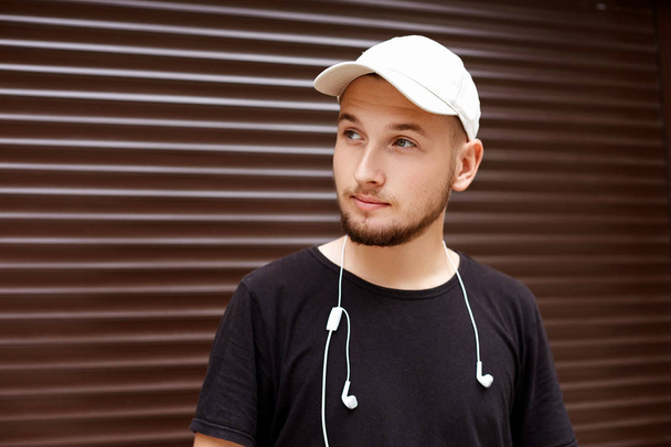 Handsome guy in a white cap and black t-shirt with headphones on the street - Zdjęcie, obraz