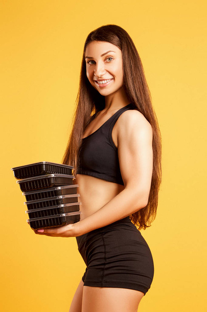 Attractive athletic girl dressed in a sports top and shorts posing in the studio on a yellow background with boxes of food. Healthy eating concept, daily diet - Φωτογραφία, εικόνα