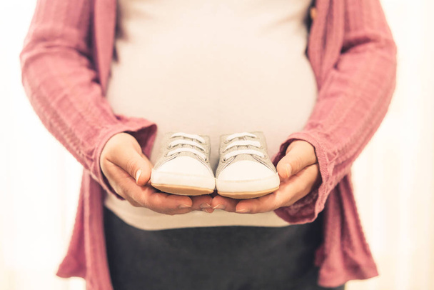 Pregnant woman feeling happy at home while taking care of child. The young expecting mother holding baby shoes. Maternity prenatal care and woman pregnancy concept. - Фото, изображение