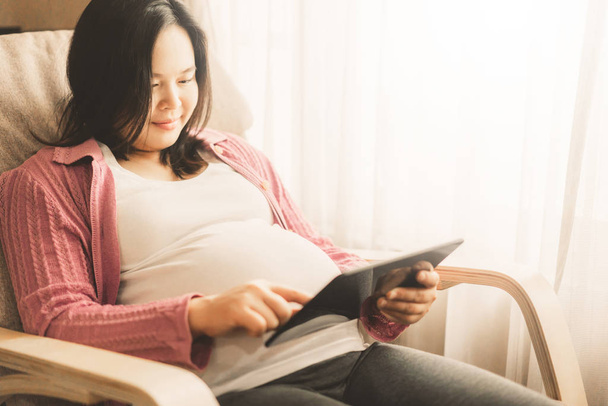 Schwangere fühlen sich zu Hause wohl, während sie sich um ihr Kind kümmern. Junge werdende Mutter sucht schwangere Fotos auf digitalem Tablet. Schwangerschaftsvorsorge und Schwangerschaftskonzept für Frauen. - Foto, Bild