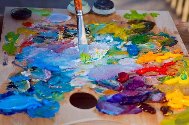 Holzpalette des Malers bedeckt mit einer Mischung aus frischen, hellen Ölfarben, Pinsel mit grüner Farbe, Unordnung beim sommerlichen Malkurs - Foto, Bild