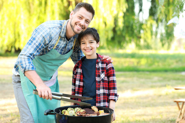 屋外のバーベキューグリルでおいしい食べ物を調理する父親と小さな男の子 - 写真・画像