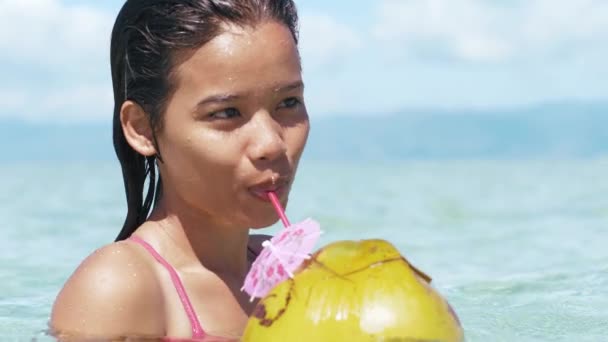 Closeup portrait of pretty asian girl in swimsuit drinks coconut on ocean beach. - Materiał filmowy, wideo