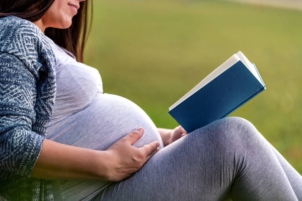 Έγκυος γυναίκα διαβάζει το βιβλίο και να χαλαρώσουν στο γρασίδι στο καλοκαιρινό πάρκο  - Φωτογραφία, εικόνα