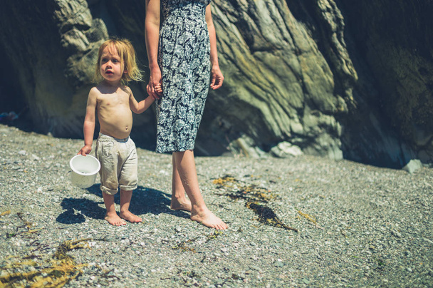 Μια νεαρή μητέρα περπατά χέρι με χέρι με το νήπιο της στην παραλία σε μια ηλιόλουστη καλοκαιρινή μέρα - Φωτογραφία, εικόνα
