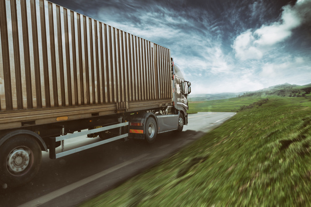 Grijze truck beweegt snel op de weg in een natuurlijk landschap met bewolkte hemel - Foto, afbeelding