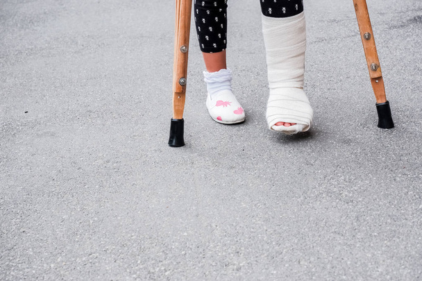 Ένα νεαρό κορίτσι σε ορθοπεδικό καστ με δεκανίκια να περπατάει στο δρόμο κοντά στο δρόμο. Παιδί με σπασμένο πόδι σε δεκανίκια, τραυματισμό στον αστράγαλο. Κάταγμα οστού και κάταγμα αστραγάλου στα παιδιά - Φωτογραφία, εικόνα