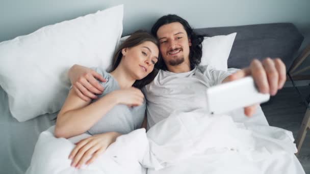 Bearded guy taking selfie in bed with girlfriend using smartphone camera - Záběry, video