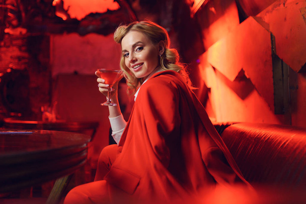 Photo of happy blonde in red jacket looking at camera with cocktail in her hand in nightclub - Foto, Imagen