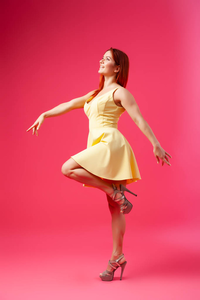Νεαρό σαγηνευτικό γυναίκα σε ένα κίτρινο φόρεμα στο ύφος της μια νέα ματιά στροβιλίζει, ποζάρει και έχει τη διασκέδαση σε ένα ροζ απομονωμένο φόντο. Η ιδέα της γυναικείας ομορφιάς και χάριτος, ποζάρισμα σε ένα φωτογραφικό στούντιο - Φωτογραφία, εικόνα