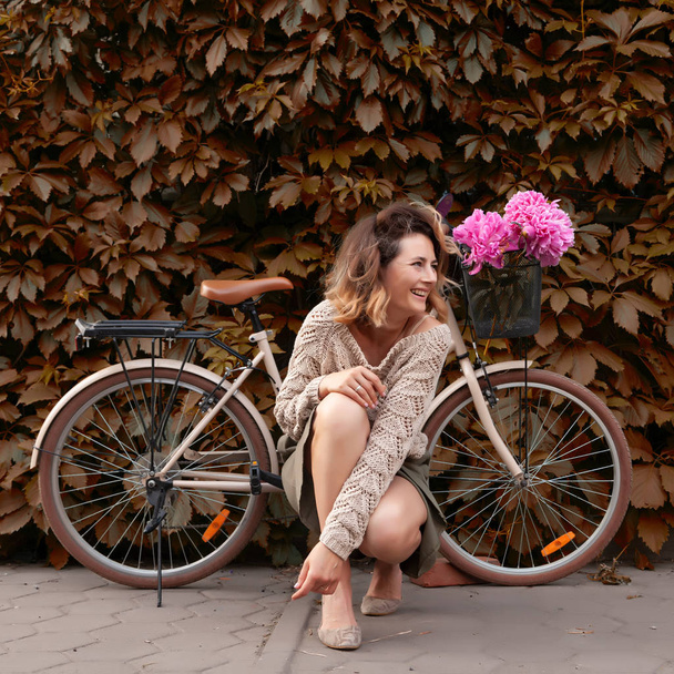 Молода жінка в жіночому одязі дивиться на камеру і позує на тлі ретро-бежевого велосипеда в літній день на фоні зеленої стіни. Концепція екологічно безпечного транспорту для жінок
 - Фото, зображення