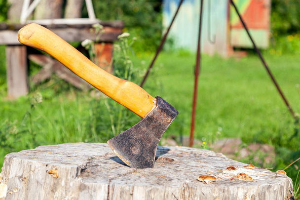 Close-up de um machado grande com uma alça de madeira presa em um toco de madeira depois de cortar lenha em um fundo verde em um dia quente de verão
 - Foto, Imagem