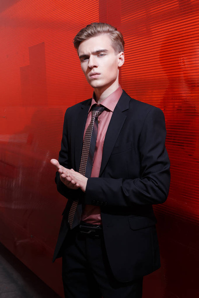 Young blond man standing and posing to the camera in the black suit on the red background - Photo, Image